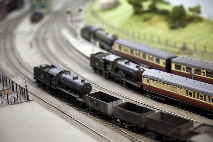 Model railway layout. A freight train with a model steam locomotive passes two passenger trains at a station. Shallow depth of field, focus on the foreground."