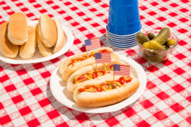 America and hot dogs. Fourth of July concept for carefree summer BBQ, unhealthy eating of junk food, Independence Day or Memorial Day backyard BBQ. Picnic food, no people. July 4th foods featured are hot dogs with ketchup and mustard and pickles. There are patriotic American flag toothpicks as decoration in the festive food. Paper plates and disposable plastic cups are also on the checkered tablecloth. Shot during sunny day outdoors on vacation. Part of a series.
