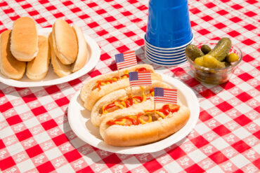 America and hot dogs. Fourth of July concept for carefree summer BBQ, unhealthy eating of junk food, Independence Day or Memorial Day backyard BBQ. Picnic food, no people. July 4th foods featured are hot dogs with ketchup and mustard and pickles. There are patriotic American flag toothpicks as decoration in the festive food. Paper plates and disposable plastic cups are also on the checkered tablecloth. Shot during sunny day outdoors on vacation. Part of a series.