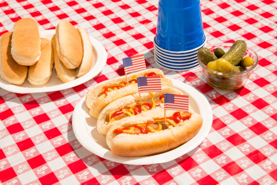 America and hot dogs. Fourth of July concept for carefree summer BBQ, unhealthy eating of junk food, Independence Day or Memorial Day backyard BBQ. Picnic food, no people. July 4th foods featured are hot dogs with ketchup and mustard and pickles. There are patriotic American flag toothpicks as decoration in the festive food. Paper plates and disposable plastic cups are also on the checkered tablecloth. Shot during sunny day outdoors on vacation. Part of a series.